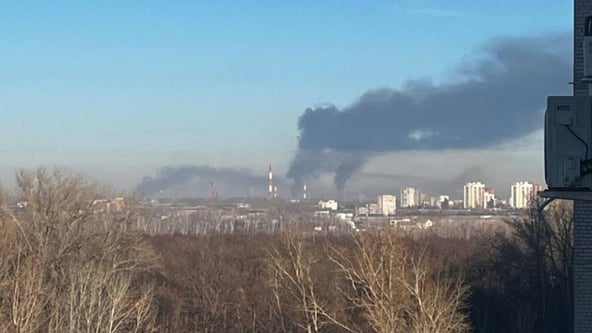 Густий чорний дим здіймається над промзоною міста на тлі безхмарного неба, на передньому плані голі дерева, вдалині житлові будинки